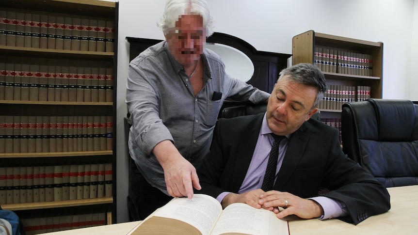 One man stands looking over the shoulder of a second man who is seated in a legal office looking at a legal book.