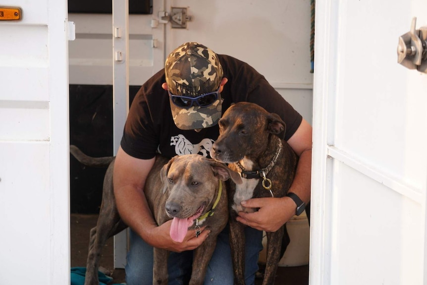 Mitch Hall wearing a cap and sunglasses on his head hugs his two dogs.