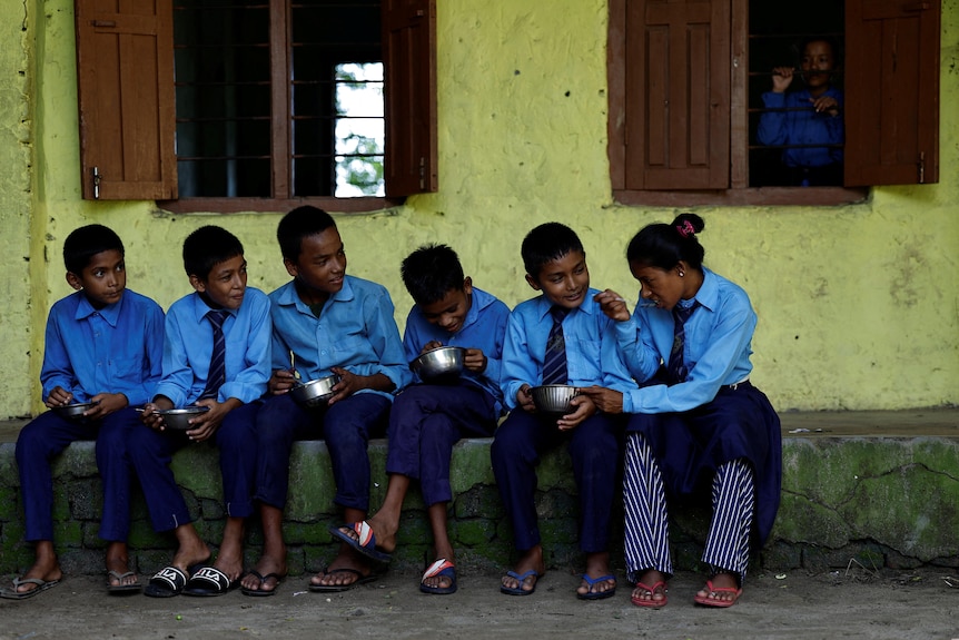 Five boys and an older woman sit on a low wall in a line, eating from silver bowls. A woman looks through a window over them