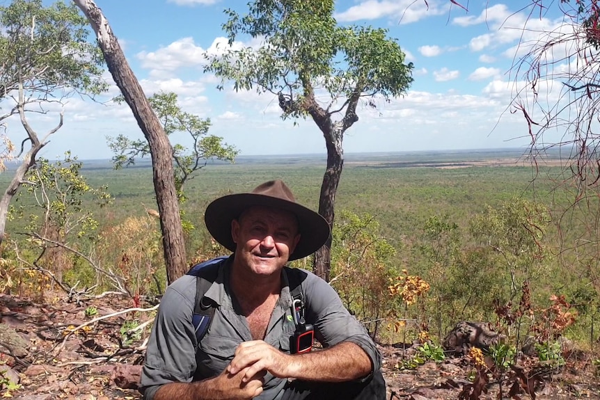 A man sits outside wearing a hat and hiking clothes. He is smiling slightly and is in the bush.