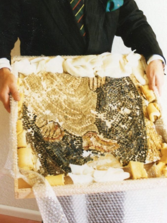 A man wearing a suit holding an ancient-looking mosaic depicting a set of feet.