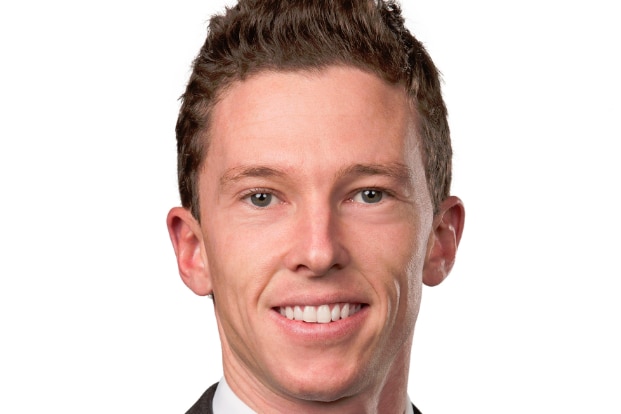 A man with brown hair smiles. He is wearing a black suit, white shirt and navy tie.