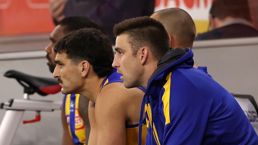 AFL players sitting on a bench, waiting to come into the game, one wearing a jacket
