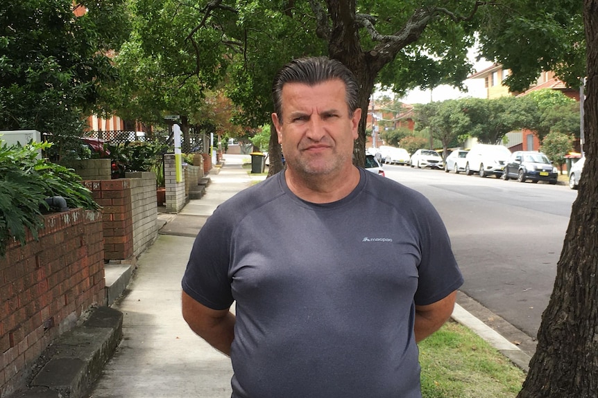 A man in a grey t-shirt stands on the footpath of a suburban street. 