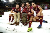 Four Queensland Origin players (three playing, one in a blazer with his arm in a sling) sit on some steps with the trophy .
