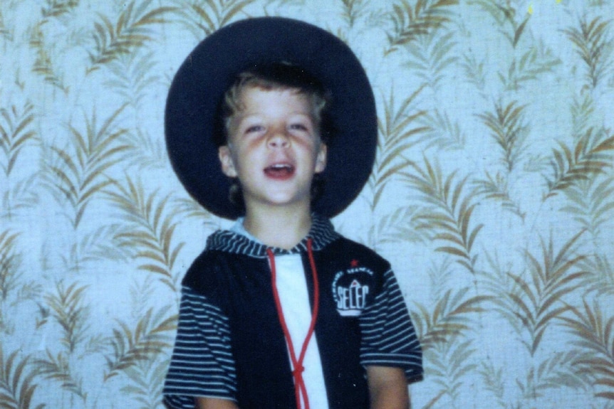A young boy, Kyle O'Neill, stands against a wallpapered wall with a sailor-style shirt on and broad-brimmed hat.