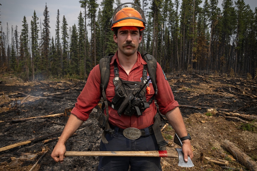 A firefighter with a moustache in a red shirt and orange helmet, strapped with equipment, holds an axe.