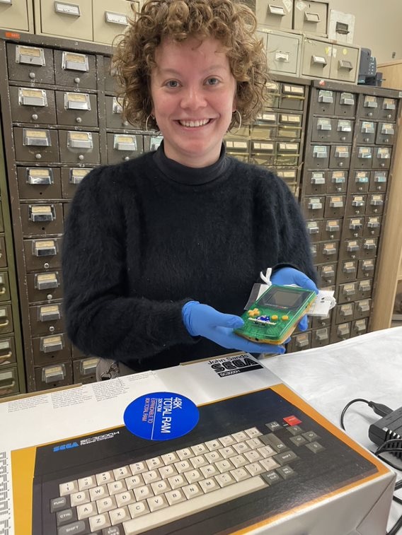 A woman smiles among old video game equipment