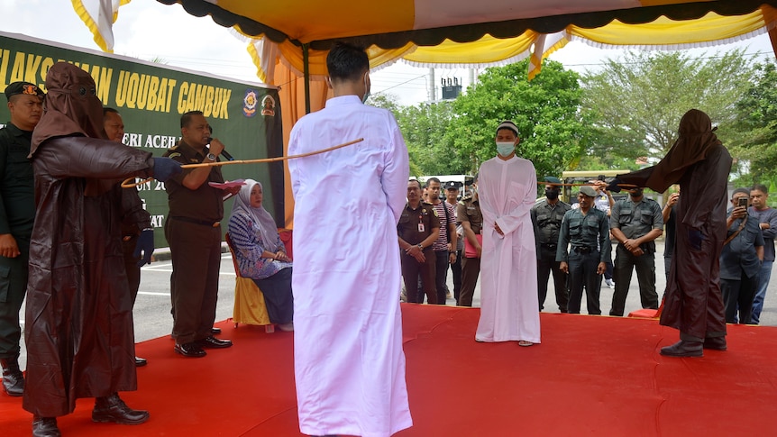 Two men in white stand and are whipped by two people dressed in black. 