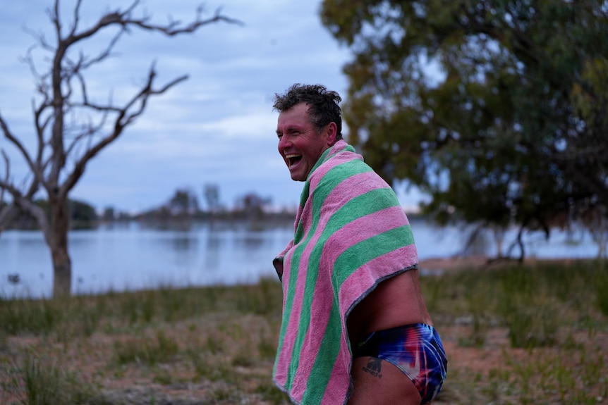A man wearing simmers and with a towel wrapped around him laughs.