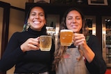 Two women with big grins hold up two glasses of beer in celebration.