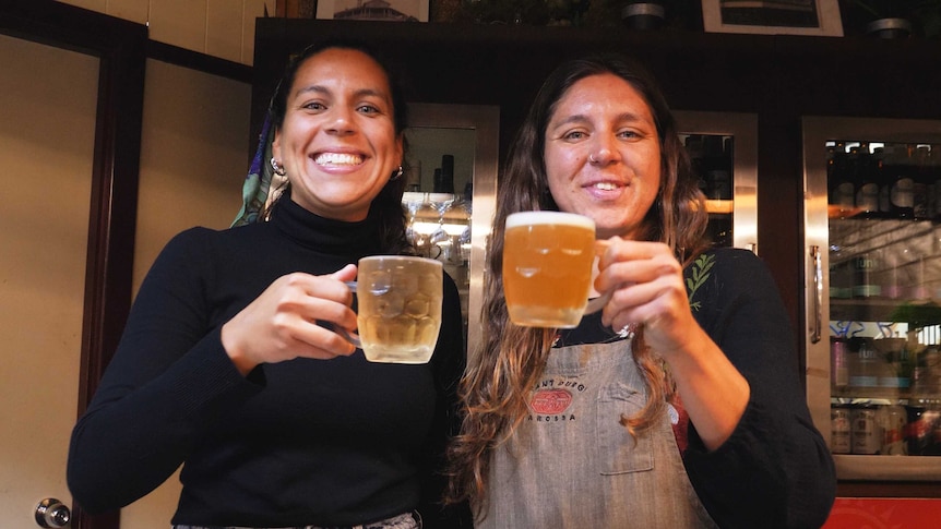 Two women with big grins hold up two glasses of beer in celebration.