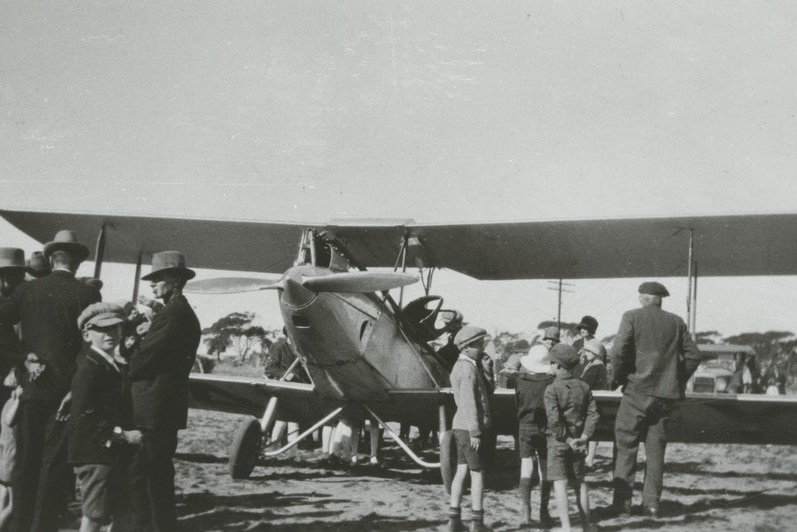 Black and white photo of people crowded in front of biplane with propeller from 1927