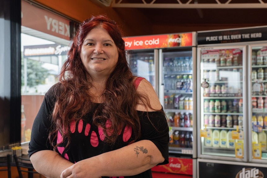 A woman with long red hair stands in front of drinks fridges in a shop