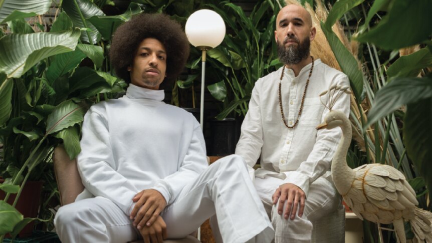 two males, one with an afro dressed in white turtleneck and jeans, the other in white linen suite with necklace