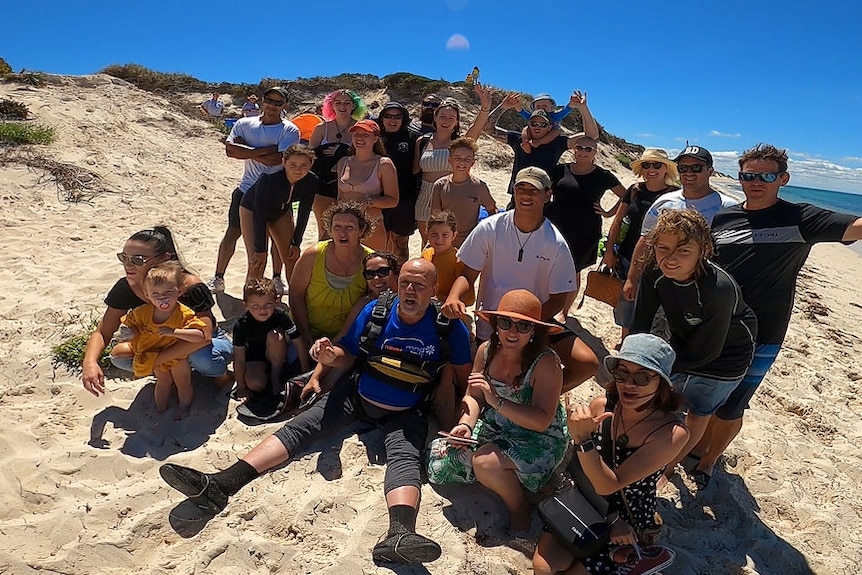 A group of people huddled together on a beach on a sunny day, smiling toward the camera.