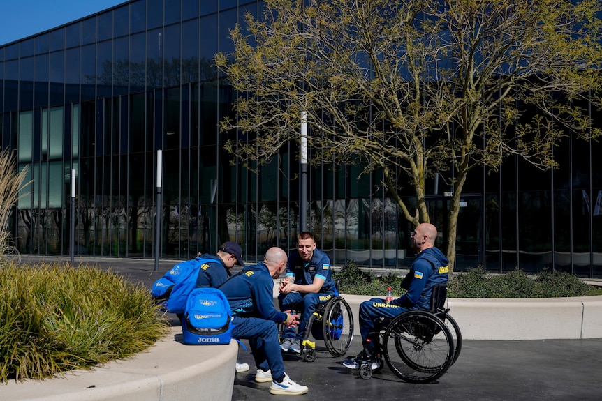 Двоє членів української команди сидять на краю грядки і розмовляють із товаришами на інвалідних візках надворі