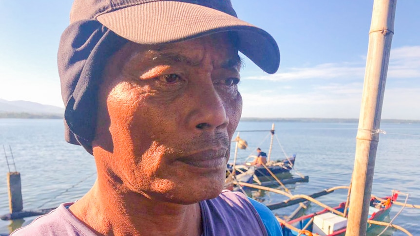 A man wearing a cap and shirt looks away from camera in front of a boat on the dock.