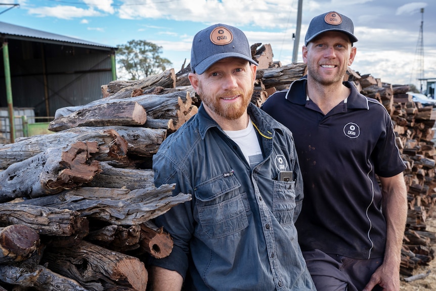 The two brothers with caps on standing in front of a pile of chopped timber.