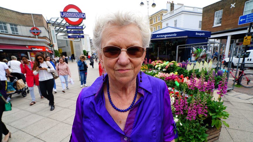 An older woman in a purple top standing next to a London tube station