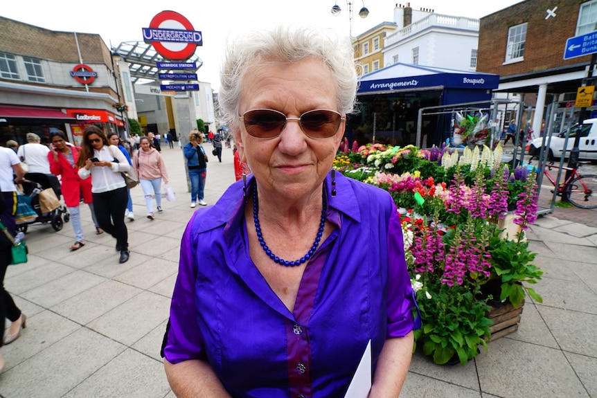 An older woman in a purple top standing next to a London tube station