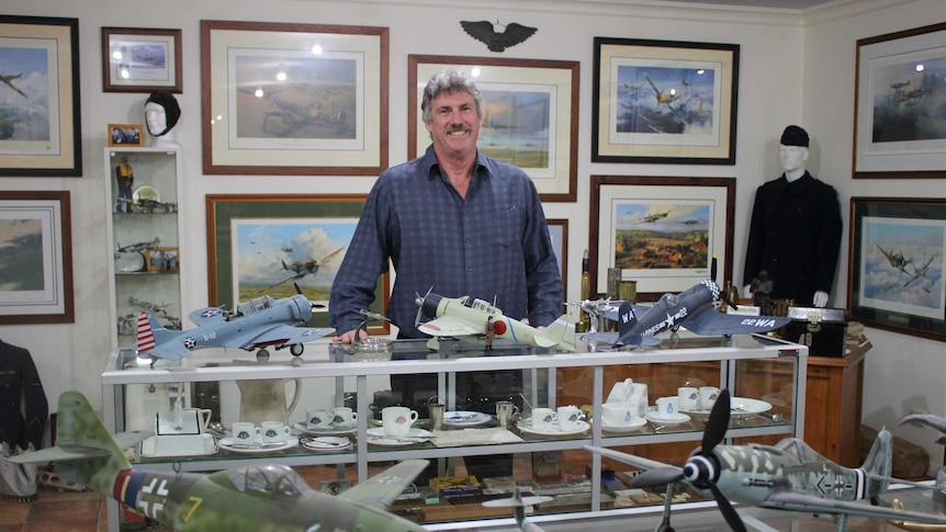 A man in a blue shirt stands in front of a museum display with aeroplane models and pictures on the walls.