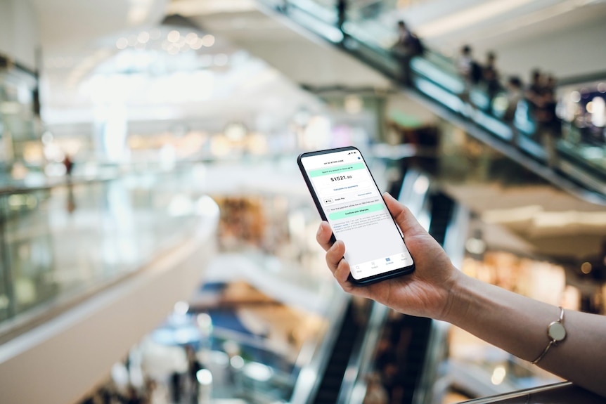 A person holds an iPhone in their right hand showing an Afterpay balance in a shopping centre with escalators in the background.