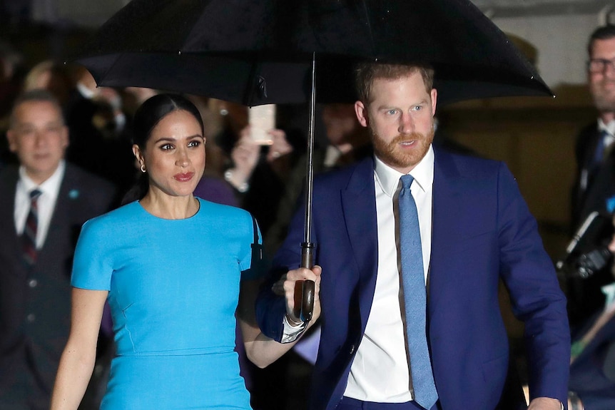 Prince Harry and Meghan, the Duke and Duchess of Sussex walk side by side holding an umbrella.