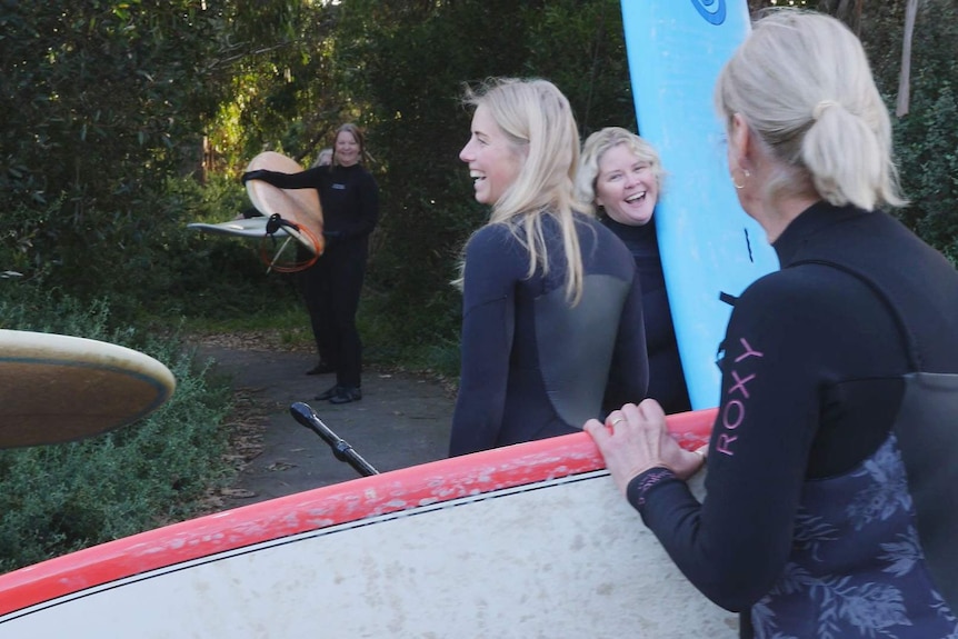 women with long-boards smiling as they head through trees to beach