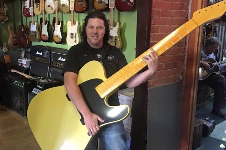 A man stands in a guitar shop holding a giant guitar