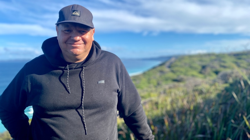 A man in a dark hoodie and cap smiles at the camera, with the ocean visible behind him