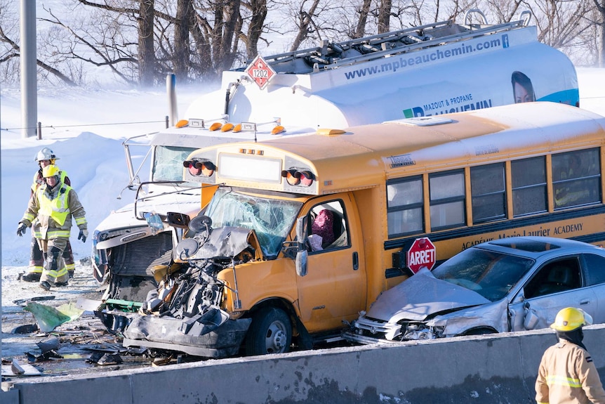 A bus rests idle with its windscreen smashed and bonnet crushed in front of a massive vehicle pileup on a snowy highway