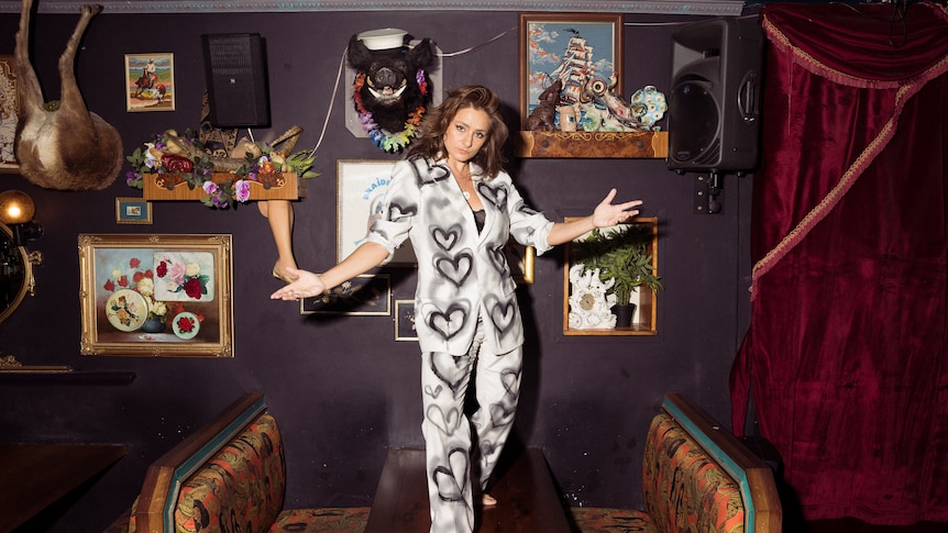 White woman with brown hair wears a white suit with spray-painted love hearts and stands on a bar table.