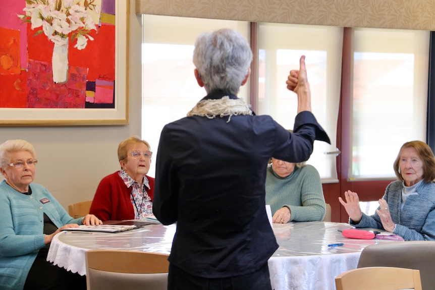 A woman with grey hair facing away from the camera gives a thumbs up.