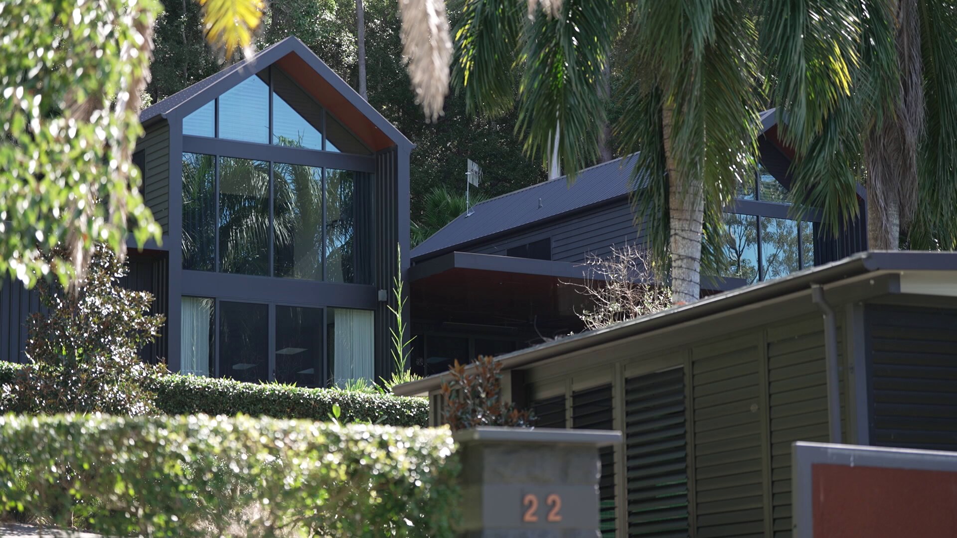A modern house with large glass windows surrounded by palm trees and other lush tropical greenery.