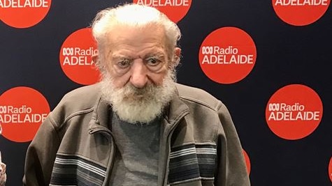 man with white hair and beard in front of black and red abc radio adelaide pul lup banner