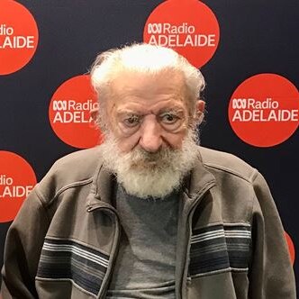 man with white hair and beard in front of black and red abc radio adelaide pul lup banner