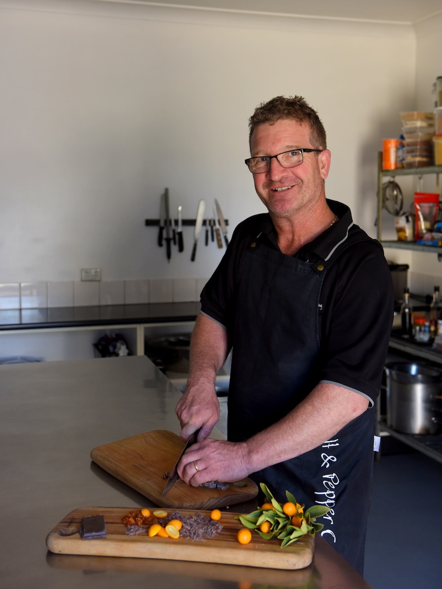 A man in a black shirt and with glasses cutting fruit on a chopping board.