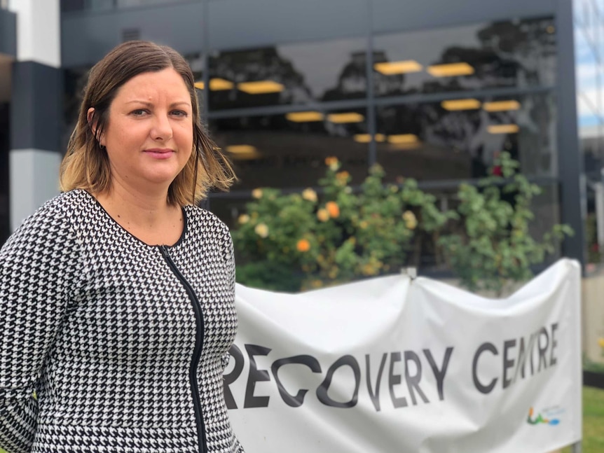 A woman in her late 30s, earing a hound's tooth jacket, stands outside near a sign reading "recovery centre".