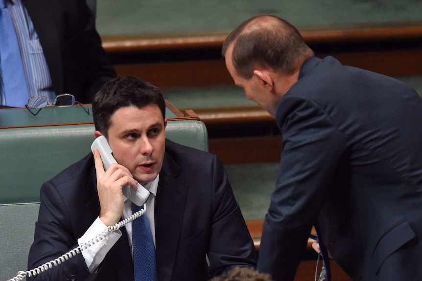 Andrew Hirst speaks on a telephone with a cord inside the House of Representatives while Tony Abbott leans over him