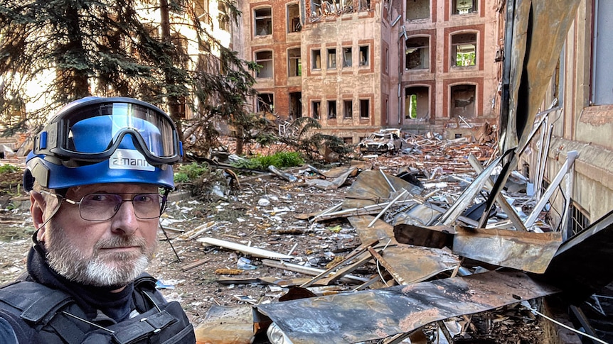 A man poses for a photo in the foreground with rubble and debris from a damaged building in the background