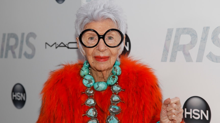 An elderly woman with white hair, dark wide glasses and red fur jacket on a red carpet.