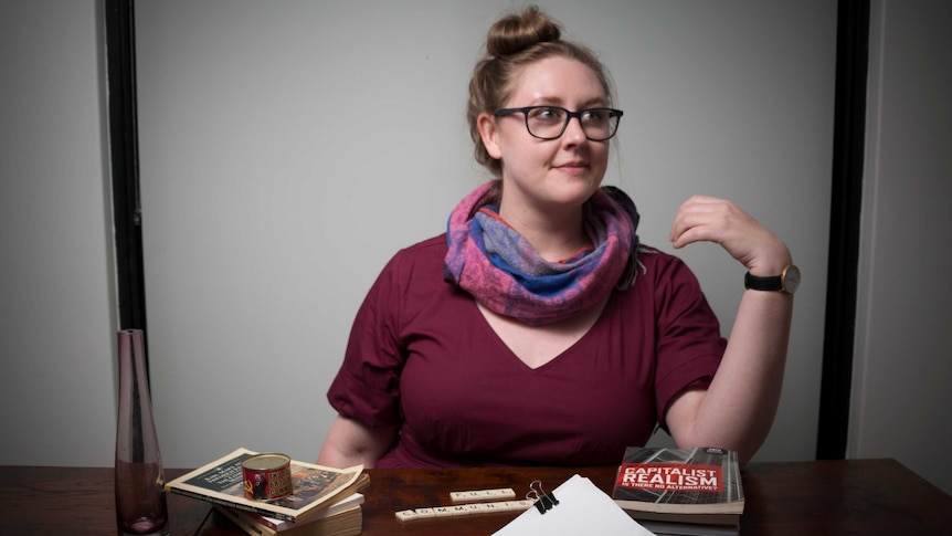 A young woman in glasses sits at a desk with left-wing literature
