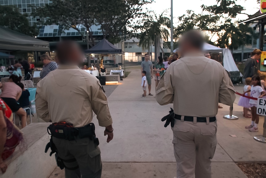 Two guards in tan khaki uniforms walk away from the camera through a market with stalls, dinders and children.