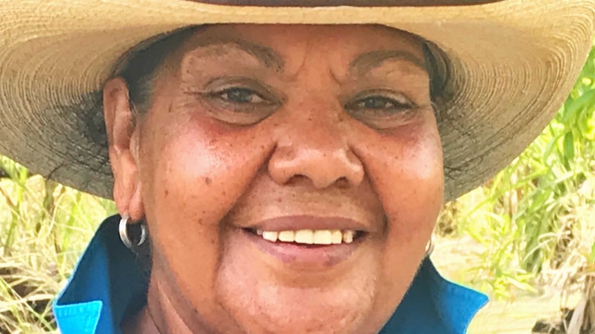 An Aboriginal woman in a straw hat smiles with lush bushland behind her.