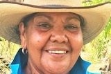 An Aboriginal woman in a straw hat smiles with lush bushland behind her.
