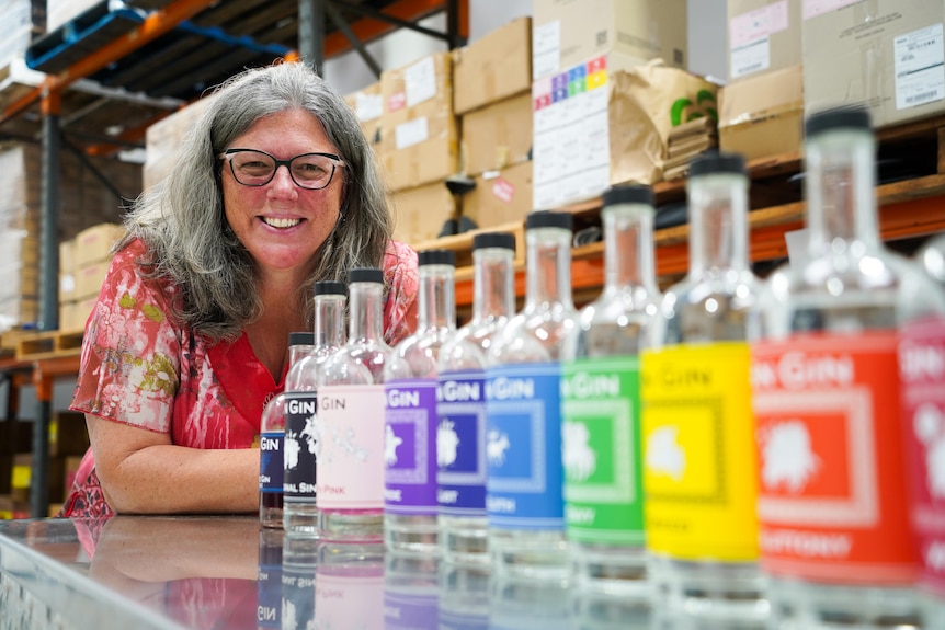 A smiling woman with grey hair and spectacles leans down on a bench behind a row of colourful gin bottles.