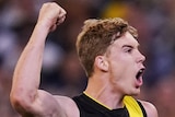 A male AFL player jumps off the ground and raises his right arm in celebration after kicking a goal.