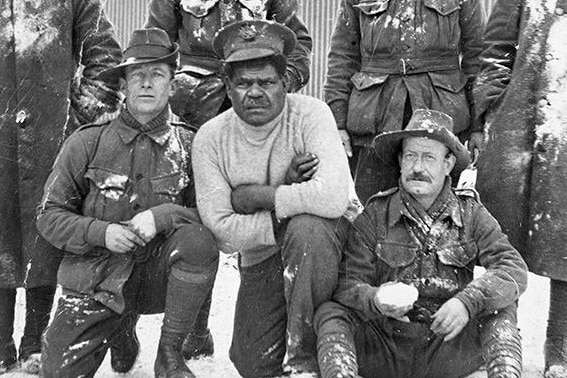 An Aboriginal man wearing a grey shirt surrounded by other men in army uniform standing in snow.