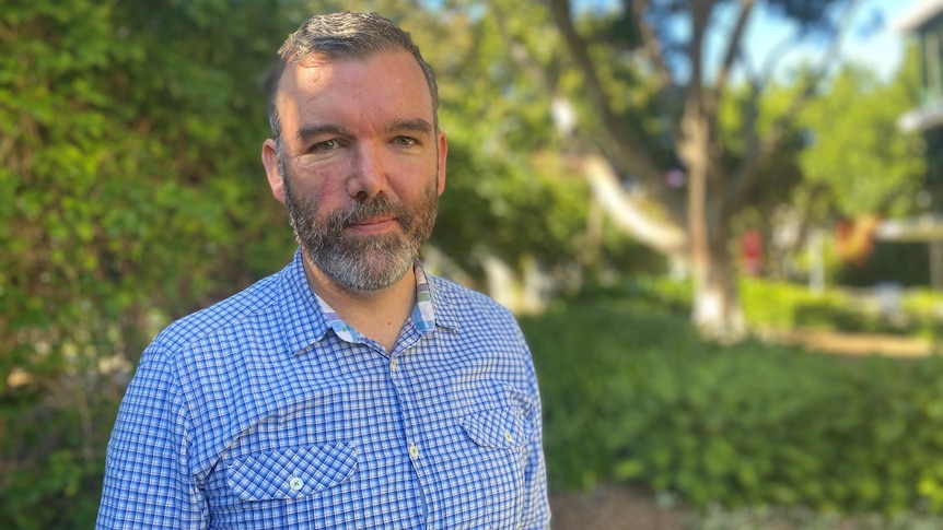 An image of Dan Angus smiling, with a blue check shirt, with blurred greenery and trees in the background